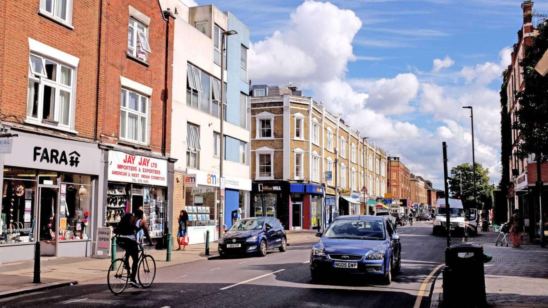 A bustling street in Bedford Hill, Wandsworth, showcasing a mix of shops, cyclists, cars, and historic brick buildings under a bright blue sky.
