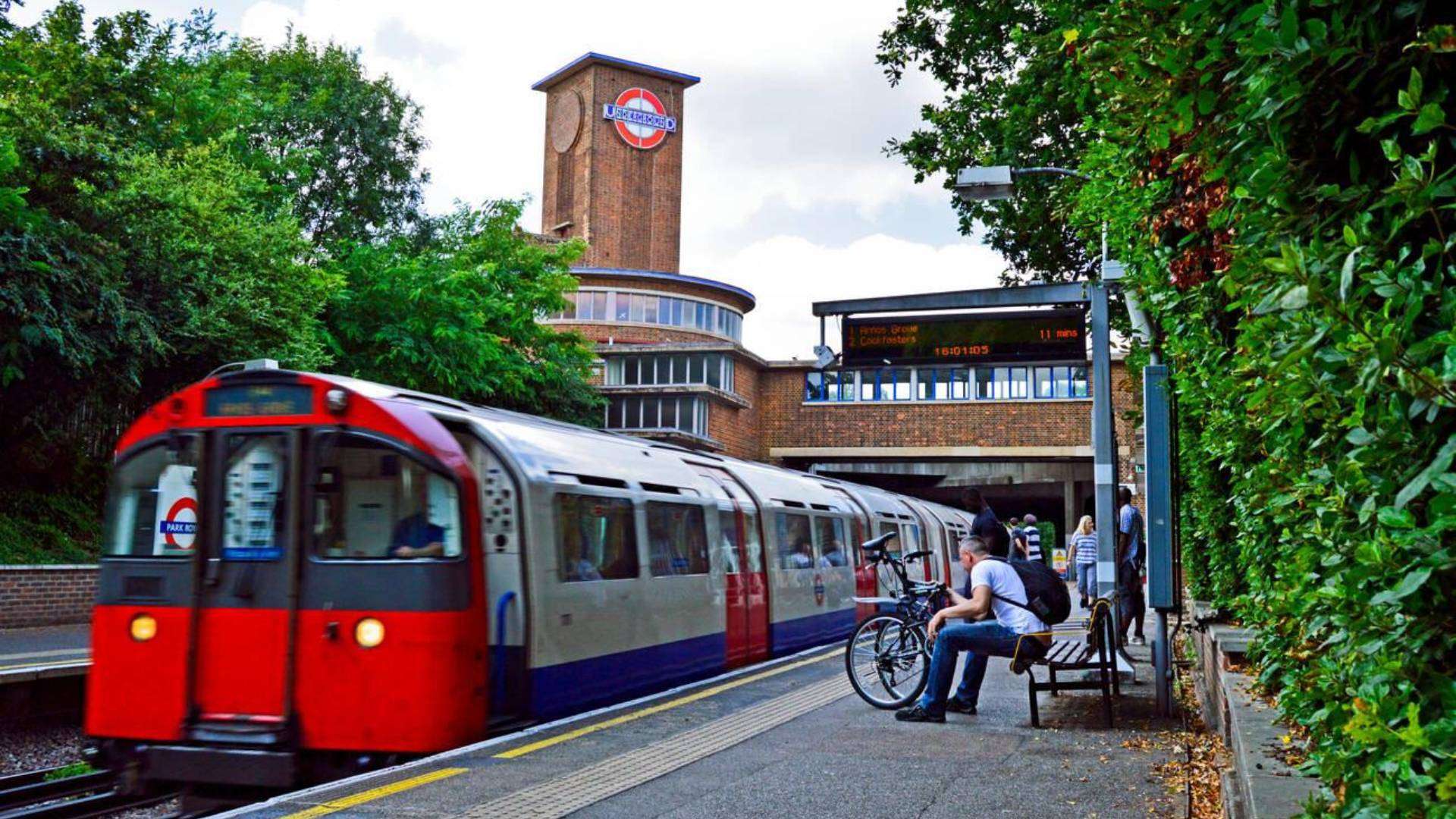 London Underground train arriving at Park Royal Station with iconic brick tower and lush greenery in the background.